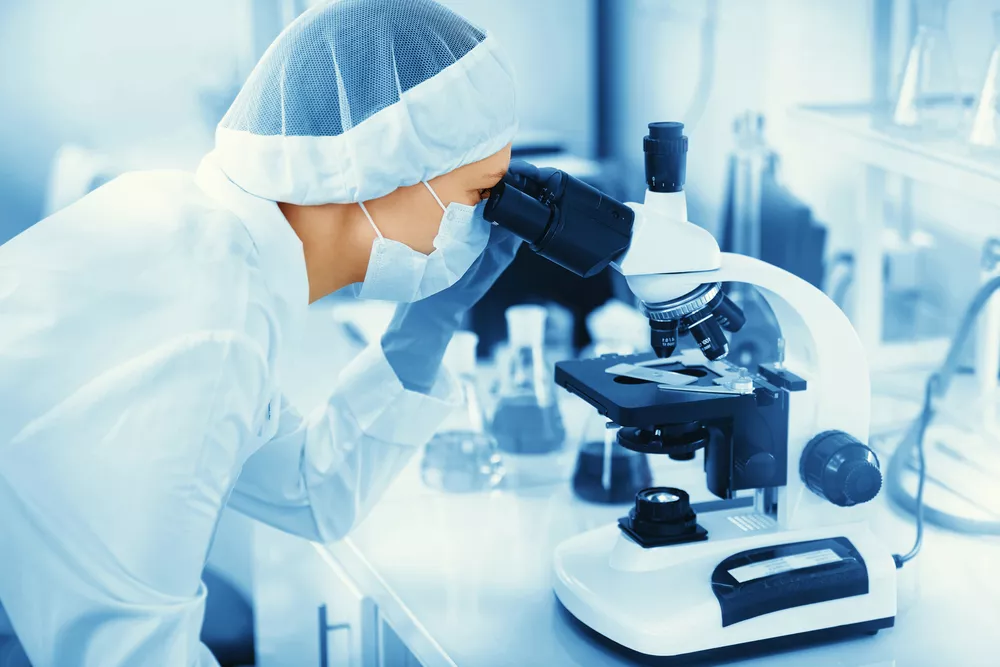 A healthcare professional wears a mask and looks through a microscope.