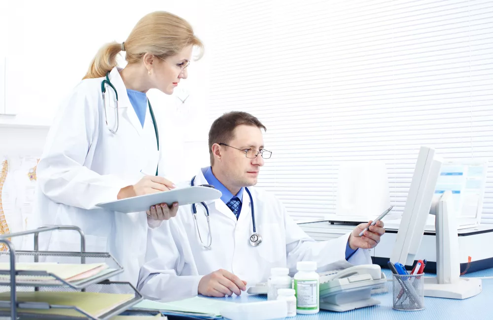 Two healthcare administrators study the screen of a computer while one writes on a clipboard.