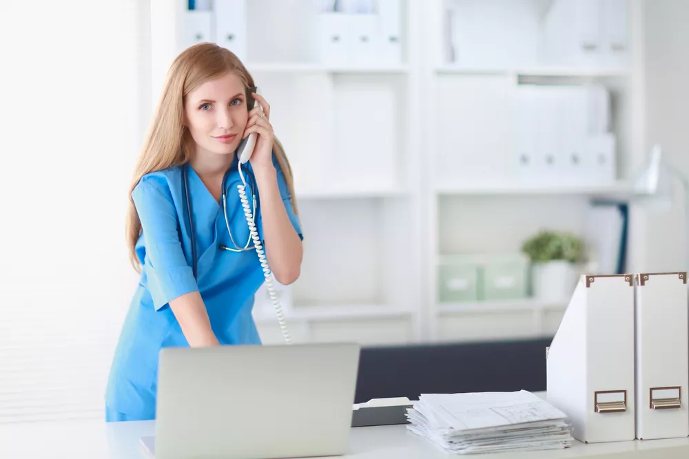 A healthcare administrator speaks on the phone while standing at a desk.