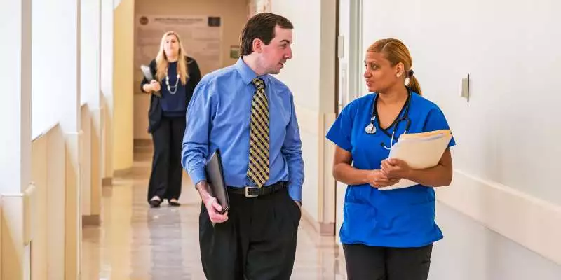 A healthcare administrator speaks to a healthcare professional as they walk down the hallway of a healthcare facility.