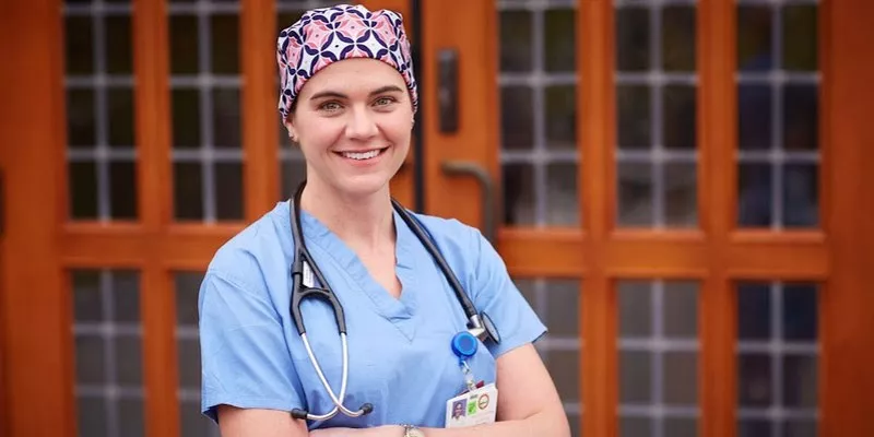 A healthcare professional in scrubs wearing a stethoscope smiles at the camera.