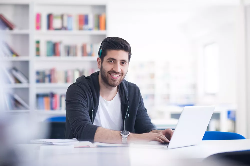 A student works at his laptop and smiles at the camera.