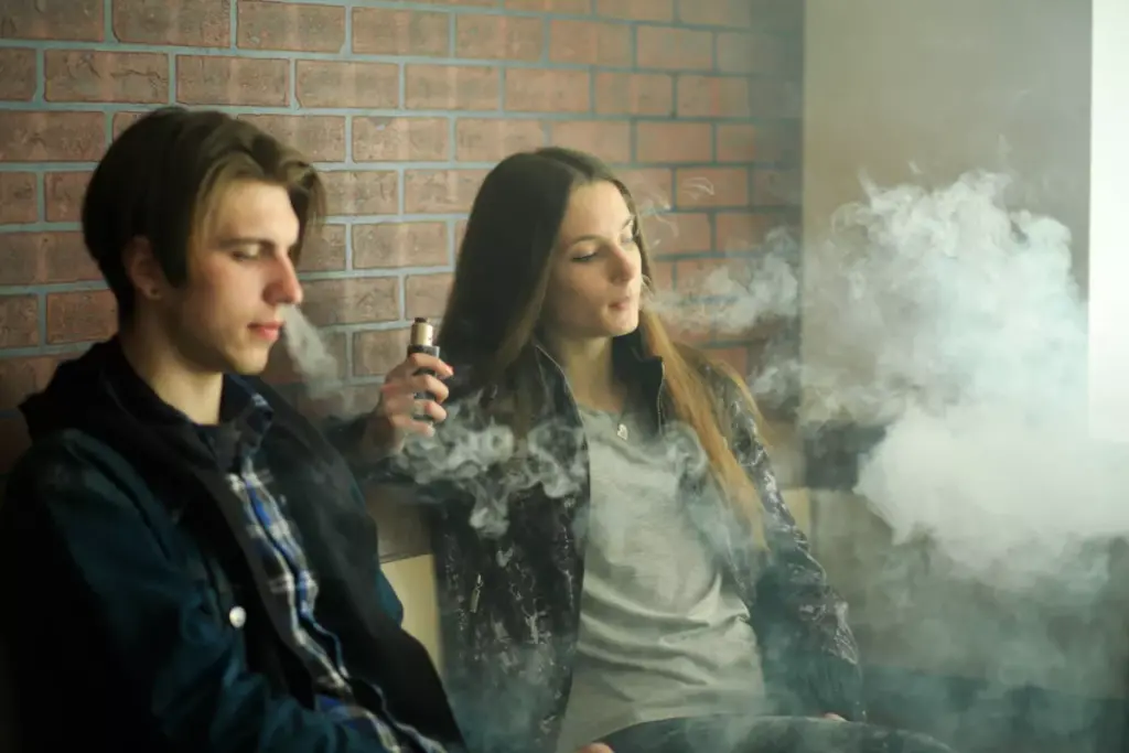 Two young adults sit side by side sharing a vape pen.