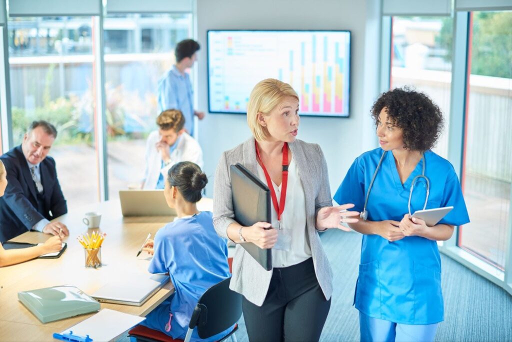 A healthcare administrator and a doctor discuss policy in a conference room; in the background, other administrators and healthcare providers confer; one studies a bar graph on a projection screen.