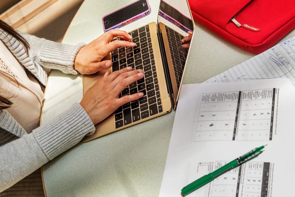 Hands typing on laptop keyboard next to smartphone, documents, notebook, and pen on table.