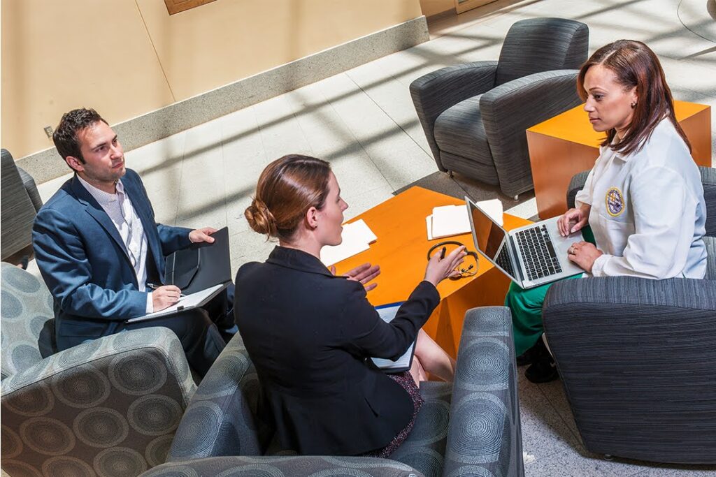Three professionals, including a woman in medical attire, sit in a sunlit lounge area having a discussion with laptops and notebooks open.
