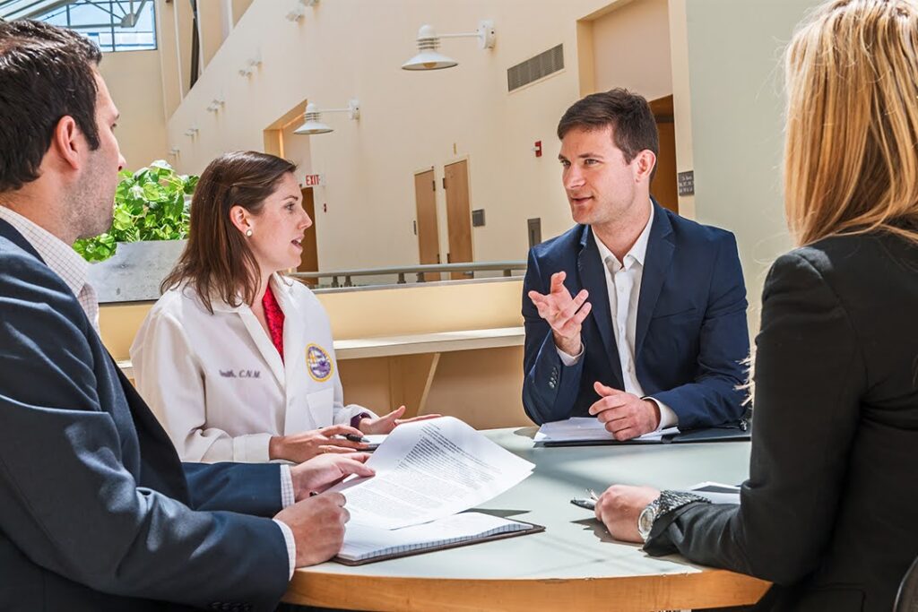 Four professionals—three in business attire and one in a white lab coat—sit around a table in discussion, with documents and notepad visible on a tabletop.