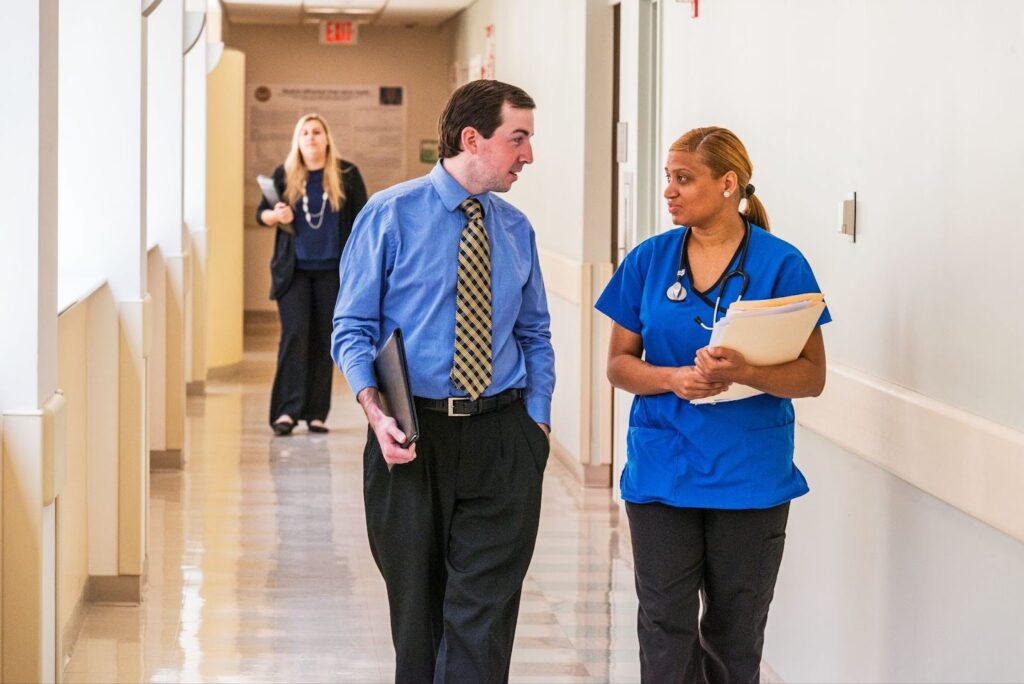 In a hospital hallway, a healthcare professional in scrubs walks and converses with a man in business attire.